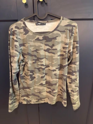 Långärmad camouflage topp - Säljer en långärmad topp med klassiskt camouflage-mönster i gröna och beige toner. Toppen har rund halsringning och är tillverkad i ett mjukt och bekvämt material, perfekt för en avslappnad och cool stil.