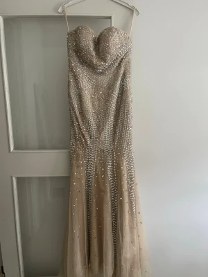 Beige axelbandslös klänning med strass - Supersnygg beige axelbandslös klänning med hjärtformad urringning och massor av glittrande strassdetaljer över hela plagget. passar perfekt för bröllop, bal och liknande fester. Klänningen har en figurnära passform upptill och faller ut i en lätt utsvängd kjol med tyll. Perfekt för dig som vill sticka ut. Pris är disskuterbart! Skickar samma dag om mäjligt går även bra att hämta och prova. Köpt i butik och använd endasy en gång! Storlek M justerbar vid bysten. Släp för mig jag är 168 cm. 