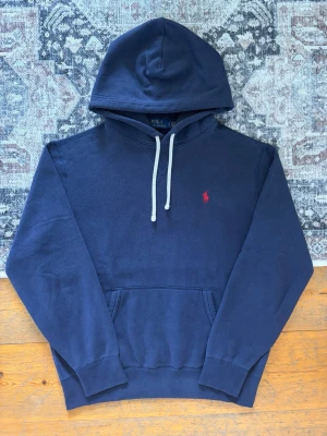 Mörkblå hoodie från Polo Ralph Lauren - Snygg mörkblå hoodie från Polo Ralph Lauren med vit dragsko och klassisk röd logga på bröstet. Hoodien har känguruficka framtill och ribbade muddar vid ärmar och nederkant. Perfekt för en avslappnad och stilren look.