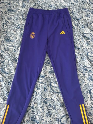 Lila Real Madrid träningsbyxor Adidas - Snygga lila träningsbyxor från Adidas med Real Madrid-logga och gula detaljer. Byxorna har smal passform, resår i midjan och gula ränder vid bensluten. Perfekta för fotbollsträning eller chill. Materialet är troligtvis polyester för maximal komfort. Inte använd 