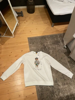Vit sweatshirt med björnmotiv - Sitter som s/m !!!! Vit sweatshirt med rund hals och ett stort tryck av en björn i streetwear på framsidan. Tröjan har långa ärmar och ribbade muddar vid ärmslut och nederkant. Perfekt för dig som gillar coola prints och avslappnad stil. Från kidsbranstore pris kan diskuteras . Kom privat för mer information 