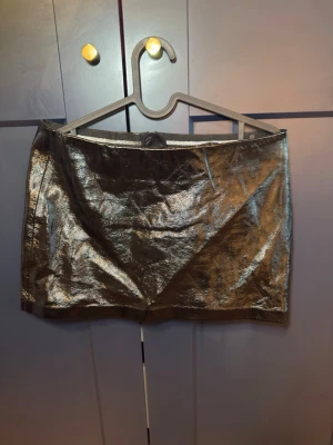 Metallic svart kortkjol i syntet - Snygg metallic svart kortkjol med glansig finish och dragkedja bak. Kjolen har en rak passform och är tillverkad i syntetmaterial som ger en cool och trendig look. Perfekt för dig som vill sticka ut med en statement piece.
