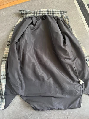 Rutig ullkappa med bälte från Lindex - Snygg lång kappa i rutigt mönster med beige, grått och svart. Kappan har knappar framtill, bröstficka med lock och ett matchande bälte i midjan. Fodrad med grått tyg för extra komfort. Perfekt för kyliga dagar och ger en trendig look.