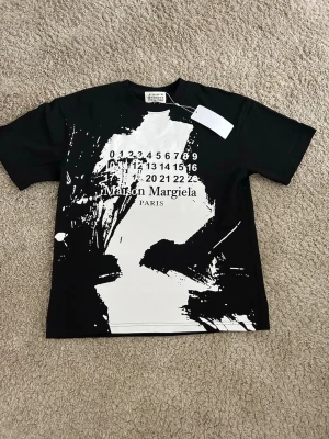Maison Margiela svart t-shirt med print - Cool svart t-shirt från Maison Margiela med unikt vitt konstnärligt print framtill och tryckt logga. Klassisk passform med rund hals och korta ärmar. Perfekt för dig som vill sticka ut med en statement-piece.