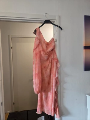Persikofärgad asymmetrisk klänning - Säljer en persikofärgad klänning med blommigt mönster och asymmetrisk design. Klänningen har en axel, lång ärm och volangdetaljer längs sidan. Midjan markeras med ett bälte och tyget är lätt och luftigt, perfekt för varma dagar.