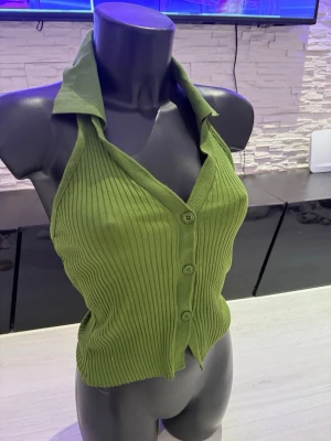 Grön ribbad halterneck topp - Snygg grön ribbad halterneck topp med krage och knappar framtill. Toppen är ärmlös och har en figurnära passform som framhäver siluetten. Materialet är stretchigt och bekvämt, perfekt för att skapa en trendig look.