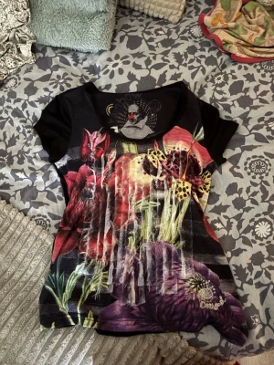 Färgstark blommig topp från Desigual - Unik topp från Desigual med färgstarkt blommönster i rött, lila, gult och grönt. Svart bas, korta ärmar och figurnära passform. Materialet känns mjukt och stretchigt, perfekt för dig som gillar statement-plagg och konstnärliga prints.