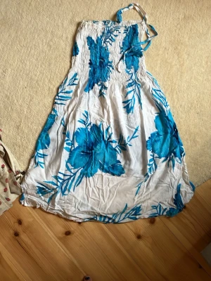 Vit långklänning med blå blommor - Säljer en somrig vit långklänning med stora blå blommor och smock upptill. Klänningen har tunna axelband och ett luftigt, svepande tyg som passar perfekt för varma dagar. Mönstret är blommigt och färgerna är klara och fräscha.