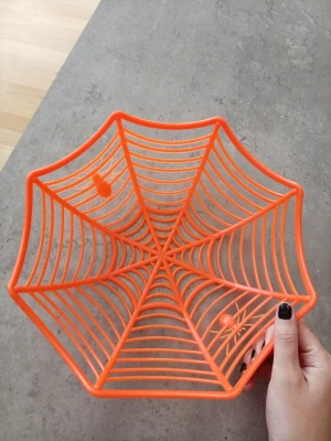 Orange spindelnäts-skål till Halloween - Cool orange skål i plast formad som ett spindelnät med små spindlar. Perfekt som dekoration eller godisskål till maskerad eller Halloweenfest. Skålen har en sexkantig form och är lätt att bära med sig.