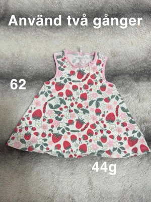 Blommig ärmlös klänning med jordgubbar - Söt ärmlös klänning i vitt med rosa kant och färgglatt mönster av jordgubbar och blommor. Klänningen har en A-linjeformad siluett och är perfekt för varma dagar. 🌸 Använd två gånger. Storlek 62 