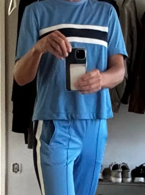 GANNI Dubois Polo Tracksuit strl S  - GANNI Dubois Polo Tracksuit, ett ikoniskt matchande set i ett mjukt, lätt och stretchigt material.  Överdel: En avslappnad, kortärmad T-shirt med tydliga, breda horisontella ränder över bröstet.  Byxor: Raka träningsbyxor med elastisk midja, kontrasterande ränder längs sidorna och diskreta dragkedjor vid anklarna. 
