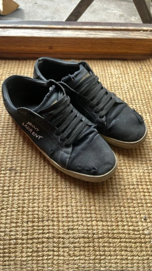 Svarta Saint Laurent sneakers - Svarta sneakers från Saint Laurent med broderad logga på sidan och gulddetaljer på plösen. Skorna har en låg silhuett, svarta skosnören och vit sula. Materialet är en mix av canvas och läder vilket ger en cool och avslappnad vibe.