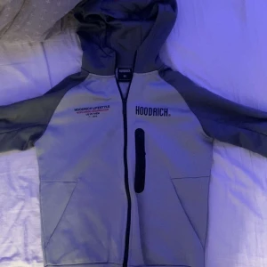 Grå zip up tröja från hoodrich - Säljer en grå zip up tröja från Hoodrich med svarta detaljer och tryck både fram och bak. Jackan har dragkedja, huva och två fickor framtill. Perfekt för dig som gillar streetwear och vill ha något stilrent men ändå sportigt. 