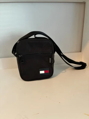 Svart axelväska Tommy Hilfiger - Snygg svart axelväska från Tommy Hilfiger med justerbar rem och dragkedja framtill. Väskan har en fyrkantig form och är dekorerad med det klassiska Hilfiger-flaggan på framsidan. Tillverkad i slitstarkt syntetmaterial.