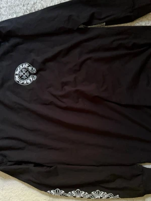 Svart långärmad t-shirt Chrome Hearts - Säljer en svart långärmad t-shirt från Chrome Hearts med vit logga på bröstet och snyggt tryck på ena ärmen. T-shirten har rund hals och är gjord i mjuk bomull som känns skön mot huden. Perfekt för dig som gillar streetwear och vill sticka ut.
