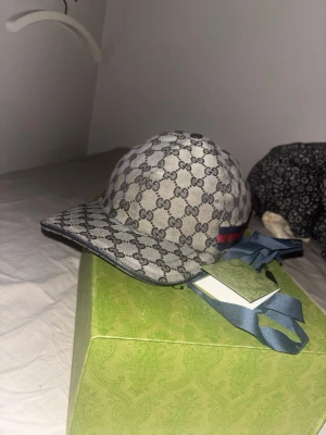 Gucci keps  - Snygg keps från Gucci i grått med klassiskt GG-monogram över hela kepsen. Kepsen har en böjd skärm och en ikonisk röd-blå rand på sidan. Materialet ser ut att vara canvas och kepsen har en modern, sportig look.