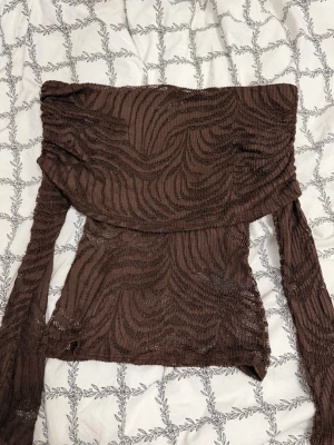 Brun offshoulder topp  - Säljer en brun offshoulder topp från H&M Edition i transparent stickad mesh med vågigt mönster. Toppen har långa ärmar och en croppad passform som ger en trendig look.  Använd 2 gånger. Som ny
