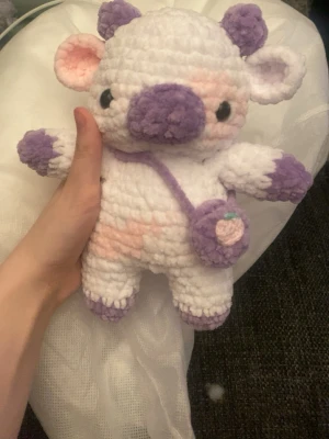 Virkad mjukisko med lila detaljer - Supergullig handvirkad mjukisko i vitt, lila och lite rosa. Den har stora öron, lila mule och små horn samt en liten axelväska i lila och rosa. Perfekt som dekoration eller present till någon som gillar söta gosedjur.