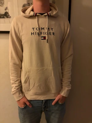 Beige hoodie från Tommy Hilfiger - Snygg beige hoodie från Tommy Hilfiger med logga och flagga broderat på bröstet. Klassisk huva med dragsnören och ribbade muddar vid ärmar och midja. Stor magficka framtill. Perfekt för en avslappnad och trendig stil.