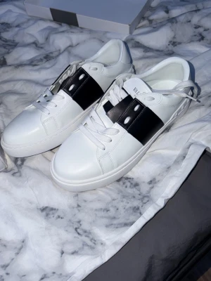 Valentino Garavani vita sneakers - Snygga vita sneakers från Valentino Garavani med bred svart läderdetalj över snörningen. Klassisk låg modell med vita skosnören och diskret logga på plösen. Tillverkade i slätt läder för en clean och modern look. Små i storlek. Nästan Oanvända
