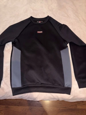 Svart sweatshirt från McKenzie - Snygg svart sweatshirt från McKenzie med grå paneler på sidorna och diskret logga på bröstet. Tröjan har rund halsringning och ribbade muddar vid ärmslut och nederkant. Perfekt för en avslappnad streetwear-look.
