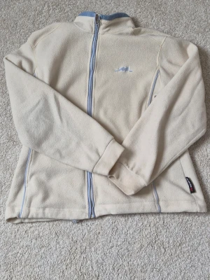 Vit fleecejacka med blå detaljer - Mysig beige fleecejacka från Tito med ljusblå dragkedja och detaljer. Jackan har hög krage, broderad logga på bröstet och två sidofickor. Perfekt för kyliga dagar och enkel att matcha med andra plagg.