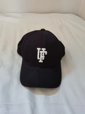 Svart keps från Upfront med broderad logga - Säljer en svart keps från Upfront med böjd skärm och justerbar snapback. Framtill sitter en vit broderad logga. Klassisk och stilren modell som passar till det mesta. Tillverkad i bomull.