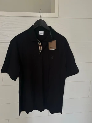 Svart pikétröja från Burberry M - Snygg svart pikétröja från Burberry med klassisk krage och korta ärmar. Tröjan har Burberrys ikoniska rutiga detalj vid knäppningen och diskret broderad logga på bröstet. Tillverkad i mjukt bomullsmaterial för en bekväm känsla.
