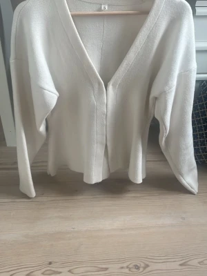 Supersöt topp - Säljer en stilren krämvit kofta med djup v-ringning och öppen framtill. Tröjan har långa ärmar och är tillverkad i ett mjukt, stickat material. Perfekt till skolan eller annan occasion🔥 den knäpps i fram enligt bilden, tröjan är figursydd med lite bredare ärmar som sitter tightare ner mot handleden