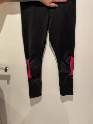 Svarta leggings med rosa detaljer - Säljer ett par svarta leggings med coola rosa detaljer vid vaderna. De har en tight passform och är perfekta för träning eller chill. Materialet känns stretchigt och bekvämt, och de har en sportig look som passar till det mesta.bak vid rumpan finns en praktisk ficka för nyckel mm.