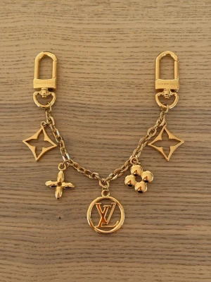 Vuitton Guldigt armband med berlocker - Snyggt armband i guldfärgad metall med flera berlocker, bland annat blommor och en rund medaljong med LV-logga. Kedjan har två karbinhakar och detaljer i form av klassiska Louis Vuitton-symboler. Perfekt accessoar för att lyfta din stil.