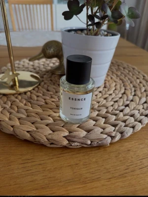 ESSNCE TOXIQUE Eau de Parfum - Minimalistisk parfymflaska från ESSNCE med doften TOXIQUE. Flaskan är genomskinlig med svart skruvkork och vit etikett med svart text. Innehållet är ljust gult och flaskan har en stilren, modern design som passar perfekt på sminkbordet.