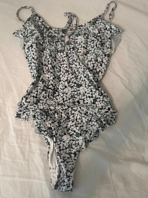 Blommig baddräkt med volanger - Söt baddräkt med svartvitt blommönster och volangdetaljer vid axlarna. Smala axelband och liten knytning framtill ger en feminin look. Perfekt för strandhäng eller poolparty. Materialet är stretchigt och bekvämt.