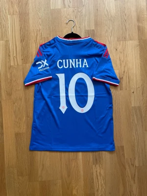 Cunha #10 - S - En liten defekt vid klubbloggan vänligen notera! Snygg blå Manchester United matchtröja med röda detaljer och vita tryck. Tröjan har CUNHA och nummer 10 på ryggen, samt klubbmärke och Snapdragon-logga på bröstet. Tillverkad i lätt och ventilerande material, perfekt för fotboll eller träning.