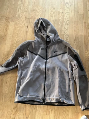 Nike tech  - Snygg grå hoodie från Nike med hel dragkedja framtill och svart logga på bröstet. Hoodien har huva, svarta detaljer och en ficka med dragkedja på ena ärmen. Perfekt för en sportig och avslappnad stil. Fin Nike tech som har används fåtal gånger.