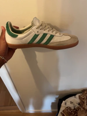 Adidas vita sneakers med gröna ränder - Säljer ett par klassiska Adidas sneakers i vitt med gröna ränder och detaljer. Skorna har en låg profil, gummisula och ovandel i läder och mocka. Perfekta för dig som gillar retrostil och sportig vibe.