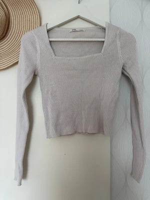 Ljusbeige ribbad topp från Sinsay - Trendig ljusbeige ribbad topp från Sinsay med lång ärm och fyrkantig ringning. Toppen är croppad och har en slim passform som framhäver figuren. Perfekt att matcha med höga jeans eller kjol för en snygg look.