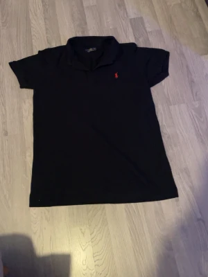 Svart pikétröja från Ralph Lauren - Klassisk svart pikétröja från Ralph Lauren med röd broderad logga på bröstet. Tröjan har korta ärmar, krage och knappar vid halsen. Perfekt till både jeans och kjol för en stilren look.