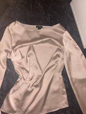 Champagnefärgad satinblus från Boohoo - Snygg champagnefärgad blus i satin från Boohoo. Blusen har långa ärmar, rund halsringning och figurnära passform med lätt markerad midja. Materialet är glansigt och ger en lyxig känsla. Perfekt att styla med jeans eller kjol. Den har en liten sminkfläck men det syns knappt❤️