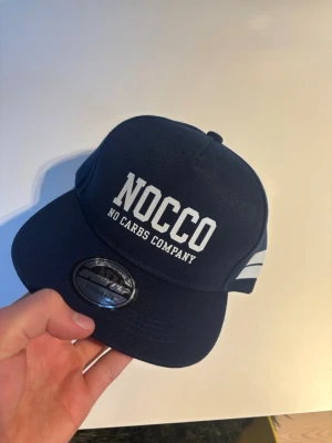 Mörkblå NOCCO snapback keps - Snygg mörkblå snapback keps från NOCCO med vit broderad text framtill och två vita ränder på sidan. Klassisk platt skärm och justerbar baksida. Perfekt för dig som gillar sportig och street-inspirerad stil.
