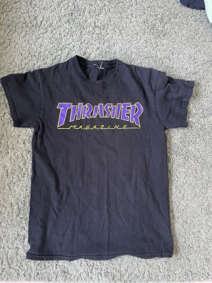 Svart Thrasher t-shirt med tryck - Säljer en svart t-shirt från Thrasher med stort tryck i lila och gult på bröstet där det står 'Thrasher Magazine'. Klassisk rund hals och korta ärmar. Perfekt för dig som gillar streetwear och skatekultur.