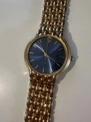 Guldfärgad klocka med blå urtavla - Säljer en elegant quartzklocka med blå sunburst-urtavla och tvåfärgat armband i guld- och silverton. Klockan fungerar som den ska och har en boettdiameter på cirka 32 mm. Diameter: 32 mm Urverk: Quartz Färg: Blå urtavla, guld- och silverfärgat armband Skick: Begagnad, fungerande Normalt slitage förekommer i form av mindre repor och användningsspår på boett och armband. Se bilder för egen bedömning. Samfrakt erbjuds vid köp av flera annonser.