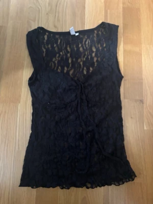 Svart spetslinne från Nelly - Säljer ett svart, ärmlöst spetslinne från Nelly i storlek XS. Linnet har v-ringning, transparent spetsmaterial och en snygg knytning framtill. Perfekt för dig som gillar trendiga och feminina detaljer.