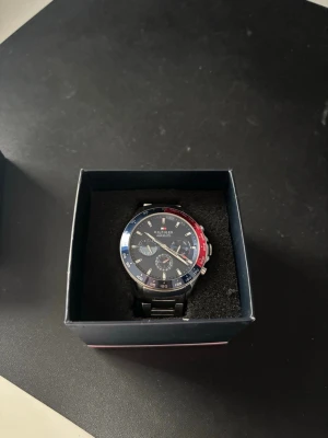 Tommy Hilfiger klocka blå/röd - Snygg klocka från Tommy Hilfiger med blå urtavla och detaljer i rött och blått på ringen. Klockan har en robust länk i silverfärgad metall och flera små urtavlor för extra funktioner. Perfekt accessoar för dig som gillar stilrena detaljer. Klockan är gjord för cirka 18-19cm passform i nuläget men finns fler länkar att addera så den blir större.