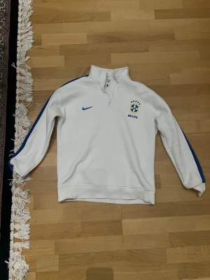 Vit Brasilien Nike träningströja - Säljer en vit träningströja från Nike med Brasilien-logga på bröstet. Tröjan har blåa detaljer längs ärmarna, halv dragkedja vid halsen och ribbade muddar. Perfekt för sport eller chill. Materialet känns mjukt och bekvämt.