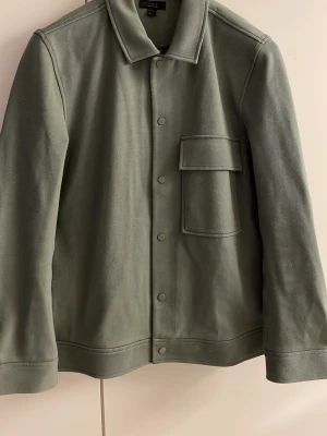 Grön overshirt med ficka COS - Stilren overshirt i dämpad grön färg med klassisk krage och tryckknappar framtill. Jackan har en stor bröstficka med lock och är tillverkad i ett kraftigare tyg med struktur. Perfekt lager-på-lager-plagg för en trendig look.