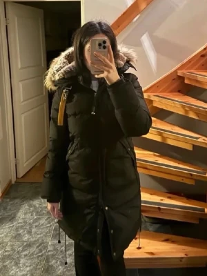 Svart lång dunjacka Parajumpers XS - Säljer en svart lång dunjacka från Parajumpers med huva och avtagbar pälskant. Jackan har dragkedja, knappar framtill och flera fickor. Klassisk gul detalj på axeln och justerbar midja för snygg passform. Perfekt för kalla vinterdagar.