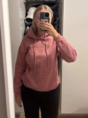 Rosa hoodie med huva och ficka - Säljer en rosa hoodie med stor magficka och dragsko i huvan. Hoodien har en avslappnad passform och ribbade muddar vid ärmar och nederkant. Perfekt för en chill och bekväm stil.
