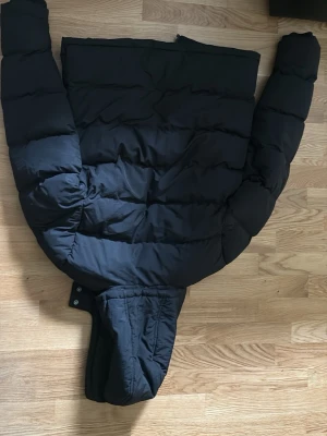 Svart pufferjacka med huva - Säljer en svart pufferjacka med huva och dragkedja framtill. Jackan har två sidofickor, quiltad design och en liten gul logga på bröstet. Perfekt för kalla dagar och riktigt skön att bära. Passar dig som vill ha en stilren och varm vinterjacka. Junior 15-16