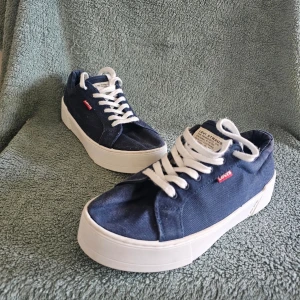 Levi's blå låga sneakers - Snygga blå sneakers från Levi's med vit tjock sula och vita skosnören. Skorna har en klassisk låg modell och är tillverkade i tygmaterial med en liten röd Levi's-logga på sidan. Perfekta för en avslappnad och trendig stil.
